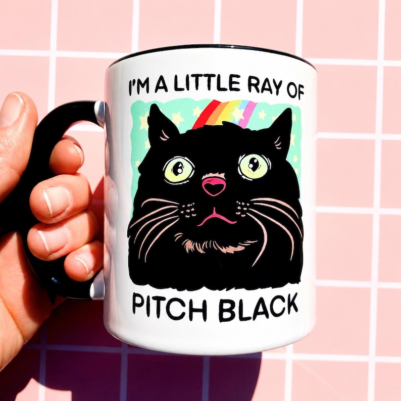 May include: White ceramic mug with a black handle and rim. The mug features a cartoon illustration of a black cat with a rainbow and the text "I'M A LITTLE RAY OF PITCH BLACK".