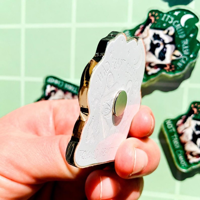 May include: A white and gold enamel pin with the text "Toward Light" and other text. The pin has a gold border and a magnetic backing. Other pins with a cow graphic and the text "It's called trash can" are in the background.
