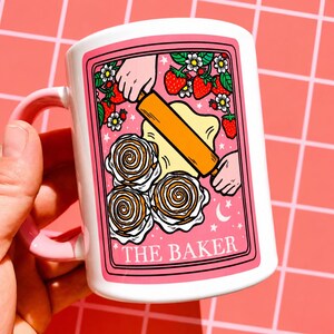 May include: White ceramic mug with a pink handle and rim. The mug has a pink framed illustration of a baker rolling dough, strawberries, flowers, and cinnamon rolls. The text "THE BAKER" is at the bottom of the illustration.