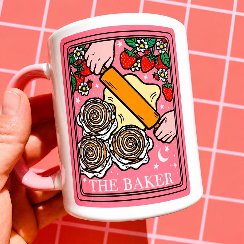 May include: White ceramic mug with a pink handle and rim. The mug has a pink framed illustration of a baker rolling dough, strawberries, flowers, and cinnamon rolls. The text "THE BAKER" is at the bottom of the illustration.