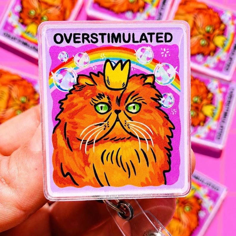 May include: A rectangular badge reel featuring a graphic of an orange fluffy cat wearing a small yellow crown. The background is purple with a colorful rainbow and floating bubbles. The text OVERSTIMULATED is printed in black at the top.