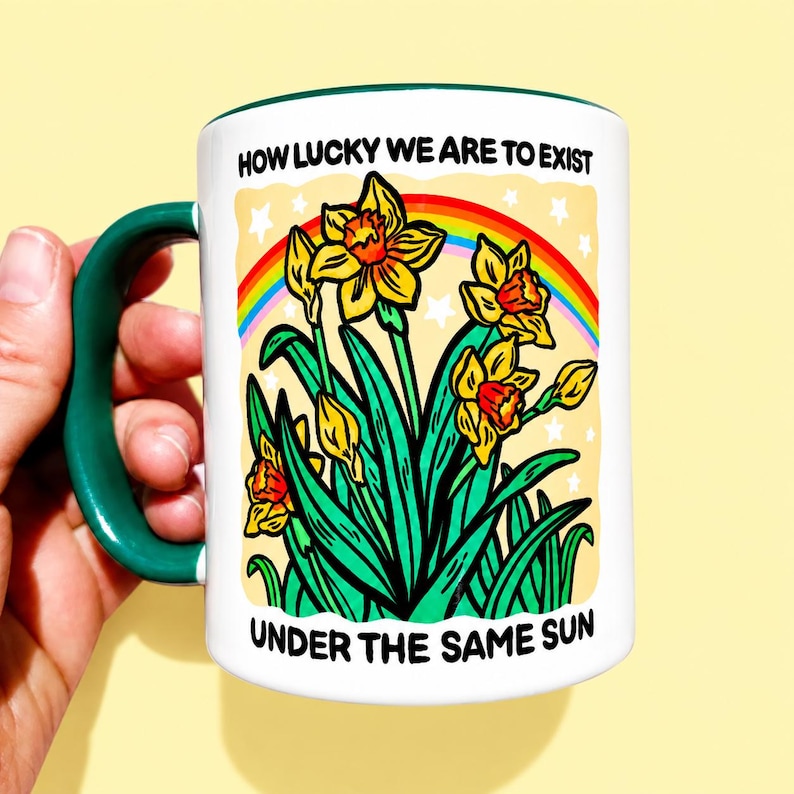 May include: A white ceramic mug with a dark green handle and rim. The front features a graphic of yellow daffodils, green leaves, and a rainbow against a pale yellow background with white stars. Text reads: HOW LUCKY WE ARE TO EXIST UNDER THE SAME SUN.