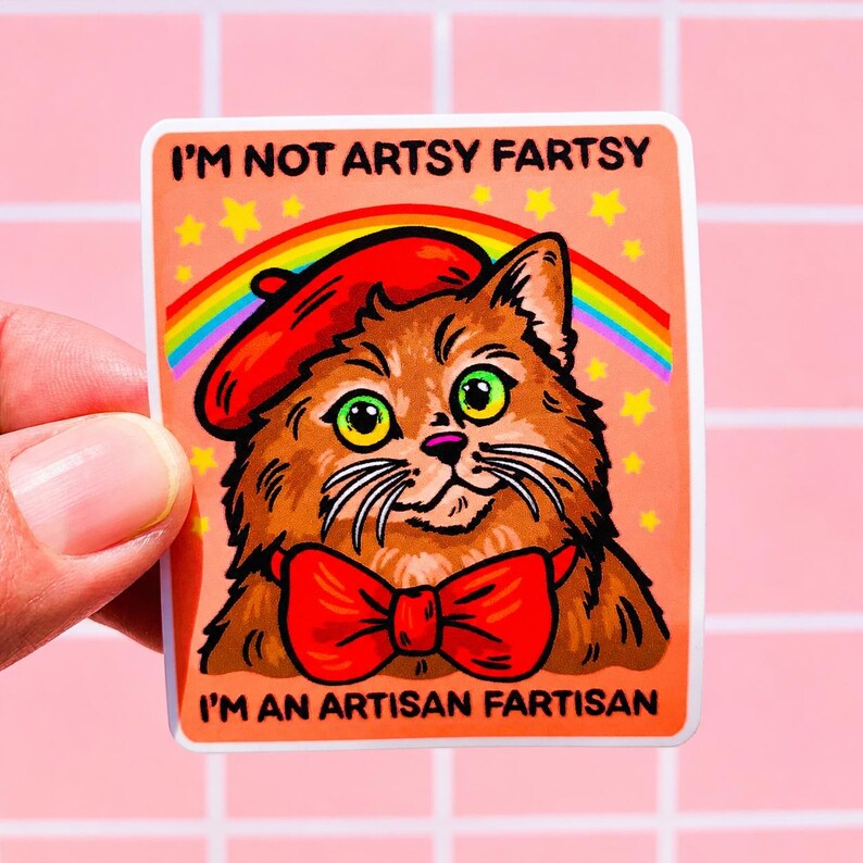 Pu&ograve; includere: Un adesivo rettangolare raffigurante un gatto marrone che indossa un basco rosso e un papillon rosso. Sopra il gatto ci sono un arcobaleno e stelle gialle su sfondo color pesca. Il testo recita I'M NOT ARTSY FARTSY e I'M AN ARTISAN FARTISAN in nero.