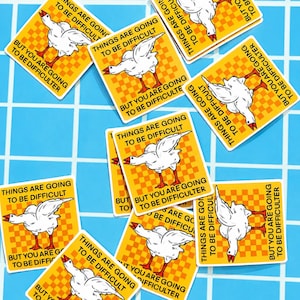 May include: A collection of yellow stickers with a white goose illustration and the text "THINGS ARE GOING TO BE DIFFICULT" and "BUT YOU ARE GOING TO BE DIFFICULTER" in black. The stickers are scattered on a blue tiled surface.