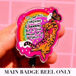 May include: A clear acrylic badge reel featuring a vibrant pink background with a rainbow, yellow stars, and a butterfly. An orange tabby cat is depicted reaching for the butterfly. The text reads: IT'S NOT ALL SUNSHINE & RAINBOWS BUT A GOOD AMOUNT OF IT ACTUALLY IS.