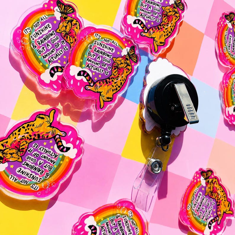 May include: Acrylic badge reels featuring a printed orange tabby cat reaching for a butterfly, a rainbow, and the text 'IT'S NOT ALL SUNSHINE & RAINBOWS BUT A GOOD AMOUNT OF IT ACTUALLY IS'. The back shows a black circular housing with a metal belt clip.