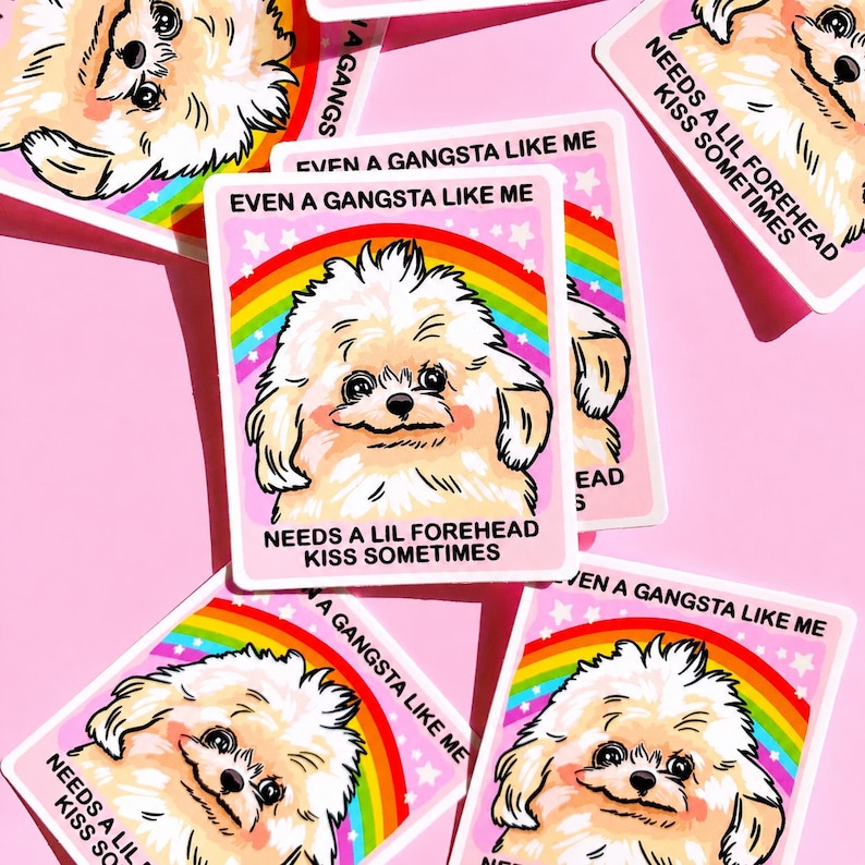 May include: A rectangular sticker featuring a fluffy cream-colored dog with a rainbow arching overhead. The text reads: EVEN A GANGSTA LIKE ME NEEDS A LIL FOREHEAD KISS SOMETIMES. The design is set against a light pink background with small white stars.