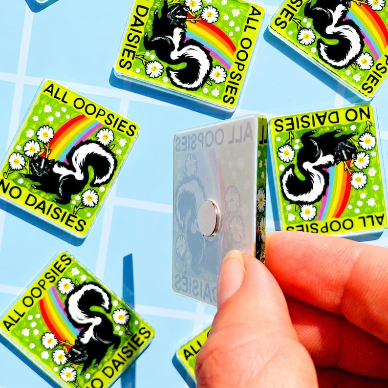 May include: A square magnet featuring a black and white skunk illustration, a colourful rainbow, and white daisy flowers on a green background. The text ALL OOPSIES NO DAISIES is printed in black. The magnet has a silver circular backing.