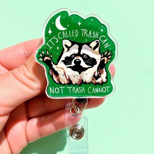 May include: A clear acrylic badge reel featuring a green background with a raccoon illustration and the text IT'S CALLED TRASH CAN NOT TRASH CANNOT. The design includes a white crescent moon and small stars, with a metal clip and retractable cord.