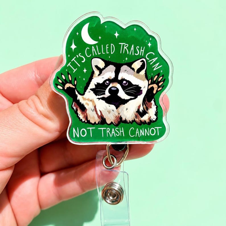 May include: A clear acrylic badge reel featuring a green background with a raccoon illustration and the text IT'S CALLED TRASH CAN NOT TRASH CANNOT. The design includes a white crescent moon and small stars, with a metal clip and retractable cord.