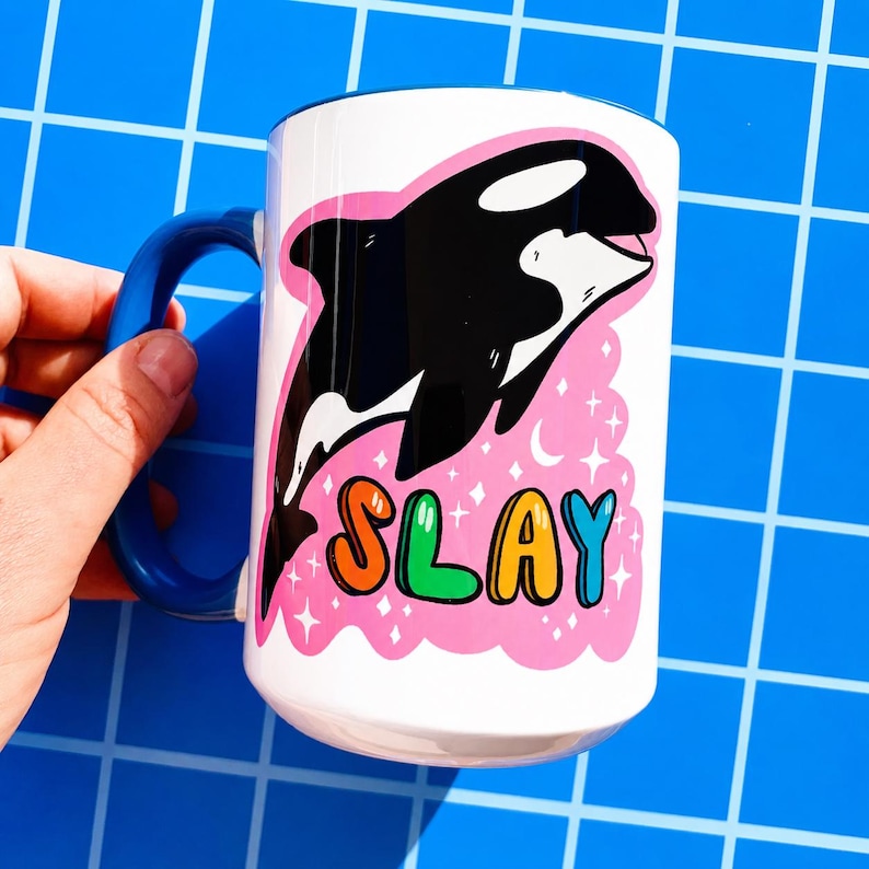 May include: White ceramic mug with a blue handle featuring a black and white orca illustration. The orca is set against a pink background with the word "SLAY" in colorful bubble letters. The mug is held against a blue grid background.