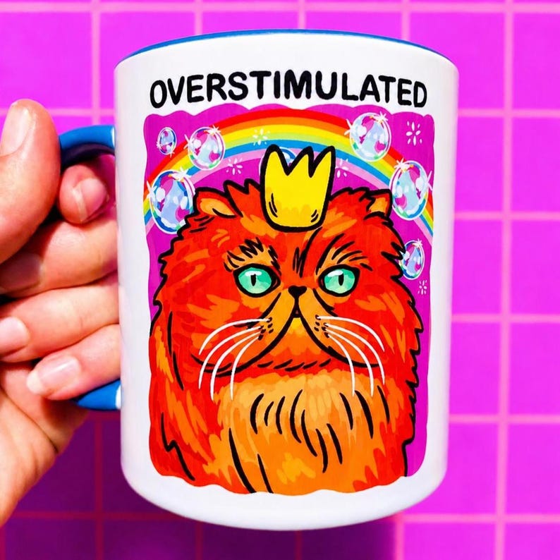 K&ouml;nnte beinhalten: Ein wei&szlig;er Keramikbecher mit blauem Henkel und Rand, der eine Grafik einer orangefarbenen Katze mit einer gelben Krone zeigt. &Uuml;ber der Katze befinden sich ein Regenbogen und Blasen, mit dem Text OVERSTIMULATED in schwarzen Gro&szlig;buchstaben.