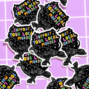 May include: A collection of stickers featuring a black crow illustration with a starry night design. Each sticker has the text "Support Your Local Murder" in colorful lettering. The stickers are on a purple grid background.