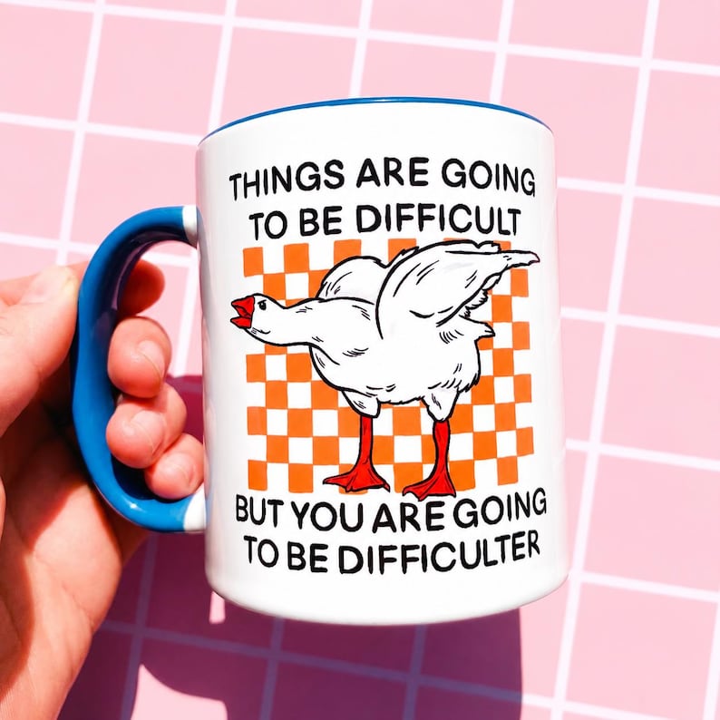 May include: White ceramic mug with a blue handle and rim. The mug features a cartoon goose illustration with the text "THINGS ARE GOING TO BE DIFFICULT BUT YOU ARE GOING TO BE DIFFICULTER" in black font. The background is an orange and white checkered pattern.