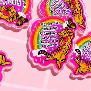 Op de afbeelding: Een doorzichtige acryl hanger met een kleurrijke illustratie van een oranje cyperse kat die naar een vlinder reikt. Het ontwerp bevat een regenboog, witte wolken en de tekst: IT'S NOT ALL SUNSHINE & RAINBOWS BUT A GOOD AMOUNT OF IT ACTUALLY IS.