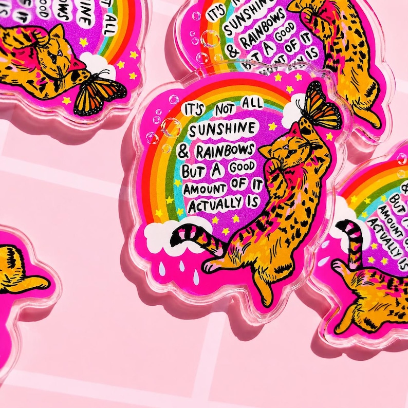 Op de afbeelding: Een doorzichtige acryl hanger met een kleurrijke illustratie van een oranje cyperse kat die naar een vlinder reikt. Het ontwerp bevat een regenboog, witte wolken en de tekst: IT'S NOT ALL SUNSHINE & RAINBOWS BUT A GOOD AMOUNT OF IT ACTUALLY IS.