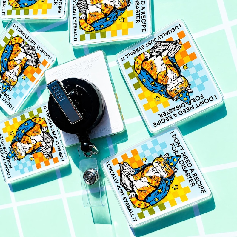 May include: Square badge reels featuring a cartoon orange cat wearing a blue wizard hat and cape, reading a book. The design includes a yellow and light blue checkered background with the text I DON'T NEED A RECIPE FOR DISASTER and I USUALLY JUST EYEBALL IT.