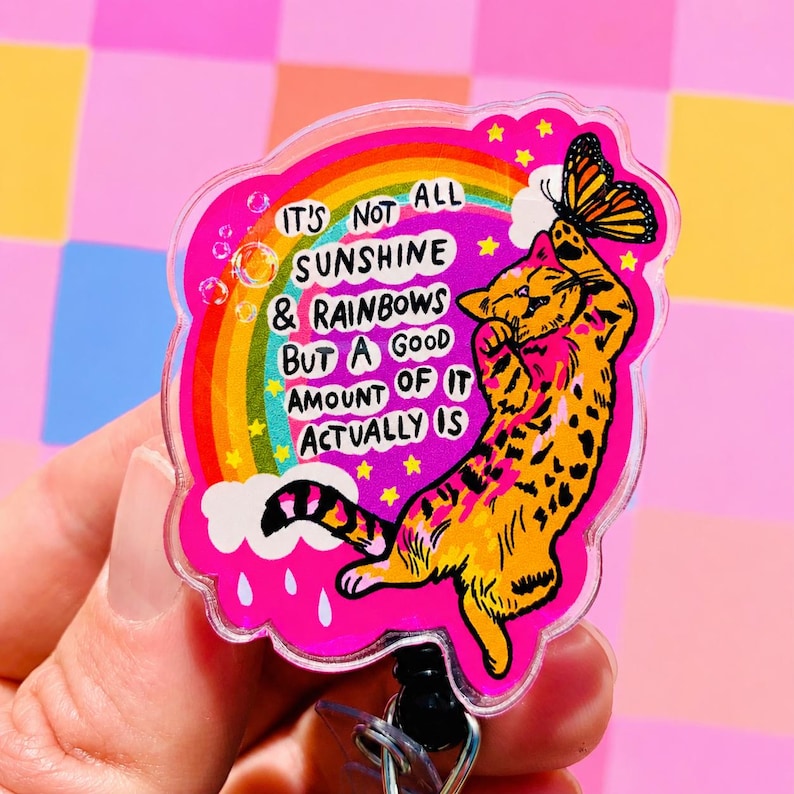 May include: A clear acrylic badge reel featuring a vibrant pink background with a colorful rainbow, white clouds, and yellow stars. An orange tabby cat reaches for a butterfly, with the text IT'S NOT ALL SUNSHINE & RAINBOWS BUT A GOOD AMOUNT OF IT ACTUALLY IS.