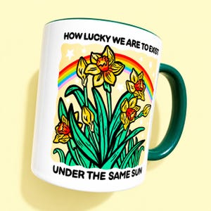 May include: A white ceramic mug with a dark green handle and interior. The front features a colorful illustration of yellow daffodils, green leaves, a rainbow, and white stars. The text reads: HOW LUCKY WE ARE TO EXIST UNDER THE SAME SUN.