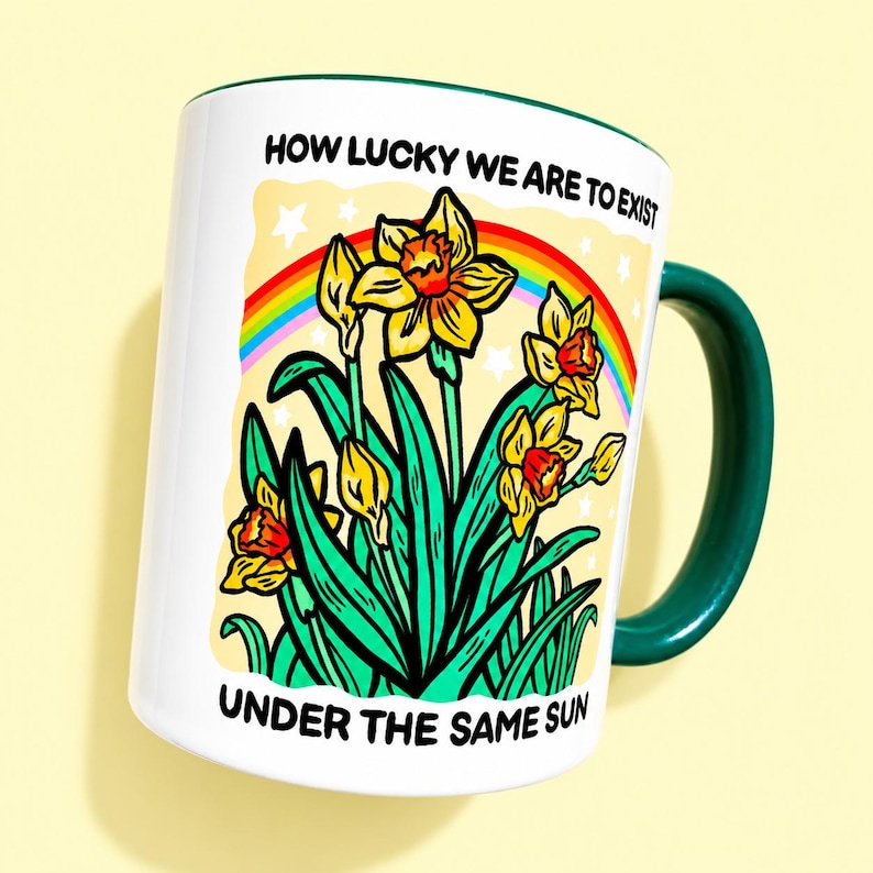 May include: A white ceramic mug with a dark green handle and interior. The front features a colorful illustration of yellow daffodils, green leaves, a rainbow, and white stars. The text reads: HOW LUCKY WE ARE TO EXIST UNDER THE SAME SUN.