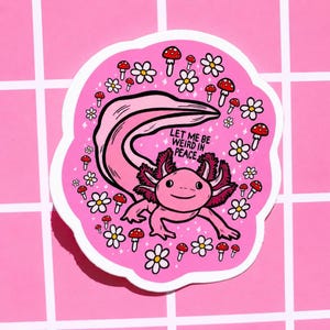 May include: A pink sticker featuring a cartoon axolotl with the text "LET ME BE WEIRD IN PEACE." The axolotl is surrounded by white daisies and red-capped mushrooms. The sticker has a white border and is on a pink grid background.