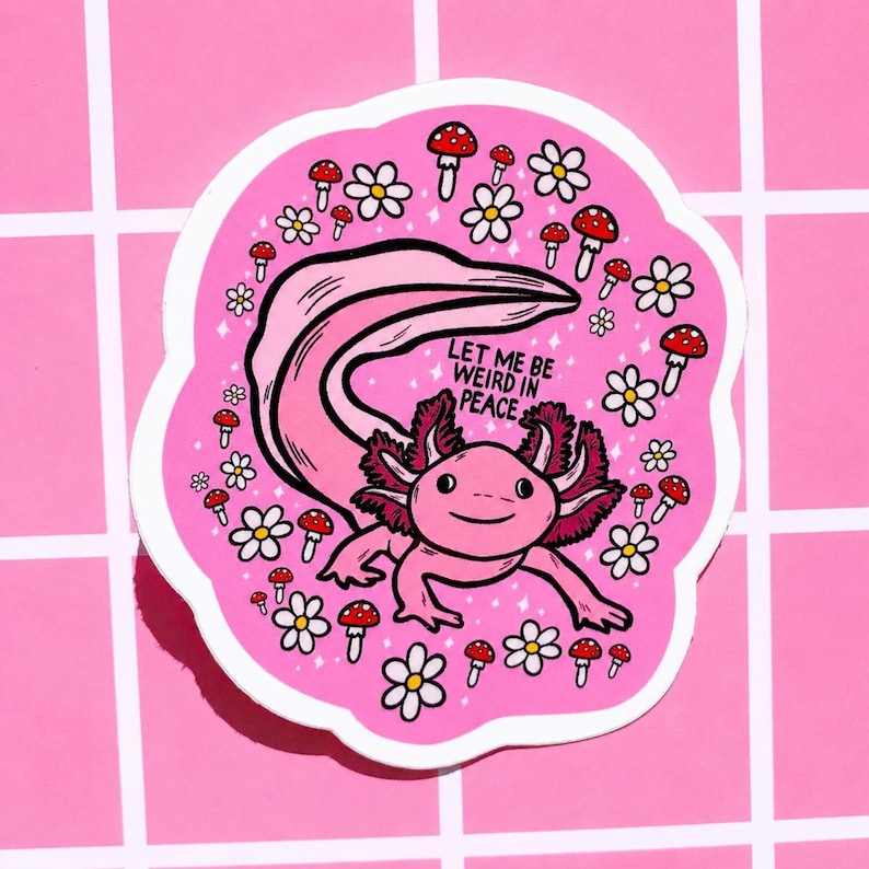May include: A pink sticker featuring a cartoon axolotl with the text "LET ME BE WEIRD IN PEACE." The axolotl is surrounded by white daisies and red-capped mushrooms. The sticker has a white border and is on a pink grid background.