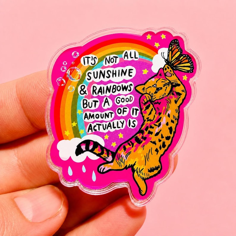 Op de afbeelding: Een gestanste acrylsticker met een felroze achtergrond, een regenboog, witte wolken en blauwe regendruppels. Een oranje cyperse kat reikt naar een monarchvlinder. De tekst luidt: IT'S NOT ALL SUNSHINE & RAINBOWS BUT A GOOD AMOUNT OF IT ACTUALLY IS.
