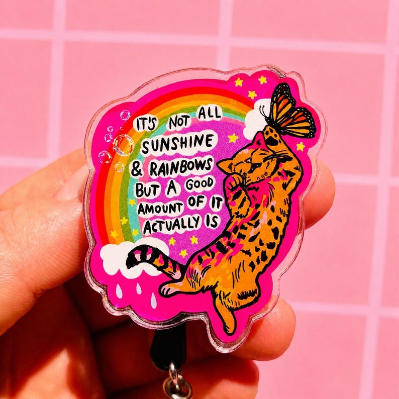 May include: An acrylic badge reel featuring an orange tabby cat reaching for a butterfly. The design includes a rainbow, white clouds, and the text: IT'S NOT ALL SUNSHINE & RAINBOWS BUT A GOOD AMOUNT OF IT ACTUALLY IS, set against a bright pink background.