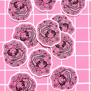 May include: A collection of pink axolotl stickers. Each sticker features a pink axolotl with a long, curved tail, surrounded by flowers and mushrooms. The text "LET ME BE WEIRD IN PEACE" is included on each sticker.
