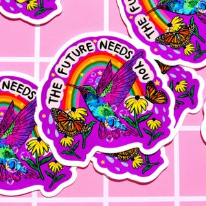 May include: A die-cut sticker featuring a vibrant hummingbird in flight, surrounded by monarch butterflies, yellow flowers, and a rainbow. The text THE FUTURE NEEDS YOU is printed in black along the top edge against a bright purple background.