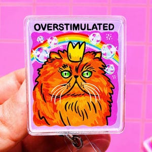 May include: A rectangular badge reel featuring a graphic of a fluffy orange cat with green eyes wearing a small yellow crown. Above the cat, the text OVERSTIMULATED is printed in black. A colorful rainbow and floating bubbles appear in the background.