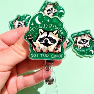 May include: A green acrylic badge reel featuring a raccoon illustration with the text IT'S CALLED TRASH CAN NOT TRASH CANNOT. The design includes a white crescent moon and stars on a dark green background, attached to a clear plastic clip.