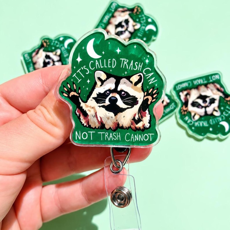 May include: A green acrylic badge reel featuring a raccoon illustration with the text IT'S CALLED TRASH CAN NOT TRASH CANNOT. The design includes a white crescent moon and stars on a dark green background, attached to a clear plastic clip.