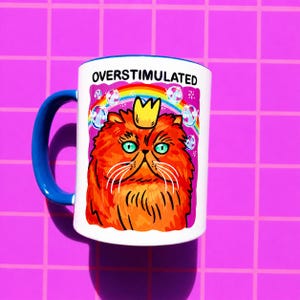 K&ouml;nnte beinhalten: Ein wei&szlig;er Keramikbecher mit blauem Henkel, der eine Grafik einer flauschigen orangefarbenen Katze mit einer gelben Krone zeigt. &Uuml;ber der Katze befinden sich ein Regenbogen und Blasen, mit dem Text OVERSTIMULATED in schwarzen Gro&szlig;buchstaben.