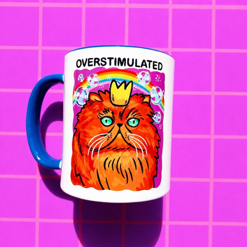 K&ouml;nnte beinhalten: Ein wei&szlig;er Keramikbecher mit blauem Henkel, der eine Grafik einer flauschigen orangefarbenen Katze mit einer gelben Krone zeigt. &Uuml;ber der Katze befinden sich ein Regenbogen und Blasen, mit dem Text OVERSTIMULATED in schwarzen Gro&szlig;buchstaben.