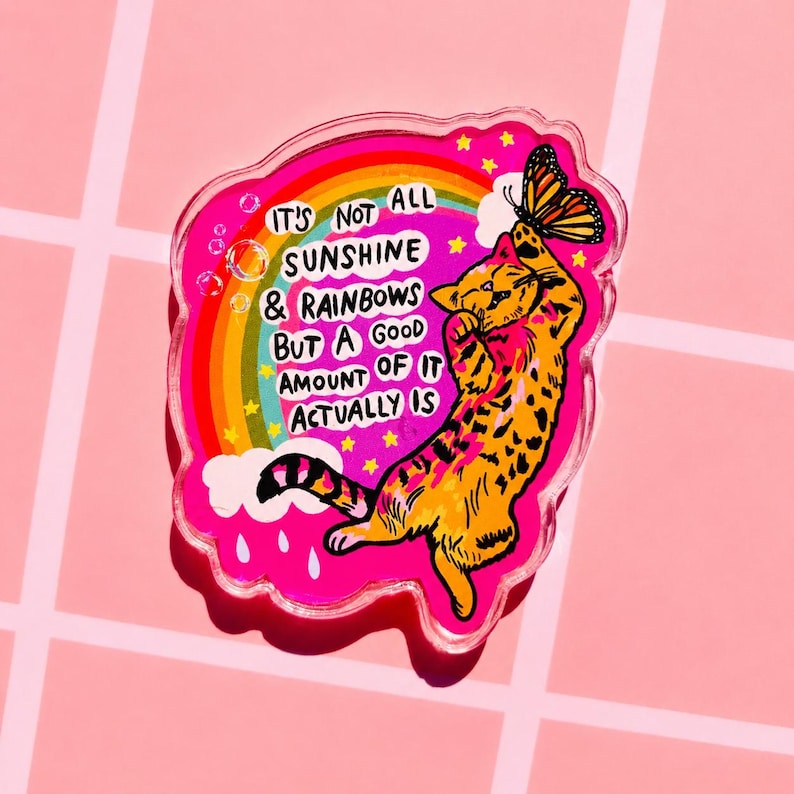 Op de afbeelding: Een doorzichtige acryl pin met een felroze achtergrond, een regenboog, witte wolken en een geel-oranje gevlekte kat die naar een vlinder reikt. De tekst luidt: IT'S NOT ALL SUNSHINE & RAINBOWS BUT A GOOD AMOUNT OF IT ACTUALLY IS.