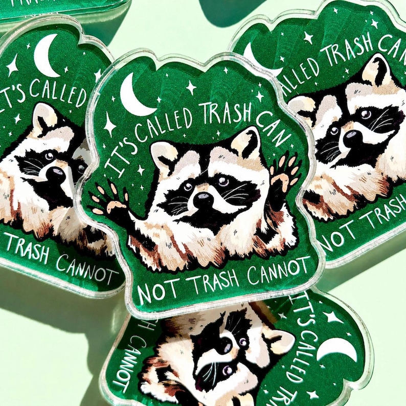 May include: A collection of green, raccoon-shaped stickers. Each sticker features a raccoon illustration with the text "IT'S CALLED TRASH CAN NOT TRASH CANNOT" in white. The background is a dark green with a crescent moon and stars.