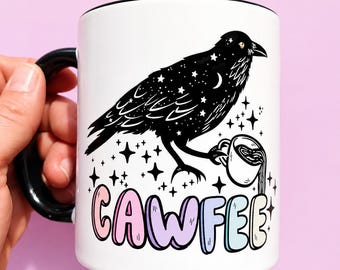 May include: A white ceramic mug with a black handle and rim, featuring a black raven illustration filled with white stars and a crescent moon. The raven holds a small white coffee cup. Below, the text CAWFEE is written in pastel-colored letters.