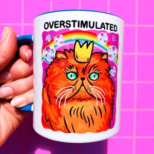 K&ouml;nnte beinhalten: Ein wei&szlig;er Keramikbecher mit blauem Henkel und Rand, der eine Grafik einer flauschigen orangefarbenen Katze mit gelber Krone zeigt. &Uuml;ber der Katze steht der Text OVERSTIMULATED. Der Hintergrund enth&auml;lt einen bunten Regenbogen und schwebende Blasen.