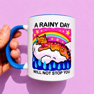 May include: A white ceramic mug with a bright blue handle featuring a colorful graphic of an orange cat resting on a cloud under a rainbow and yellow stars. The text reads A RAINY DAY WILL NOT STOP YOU in bold black letters above and below the graphic.