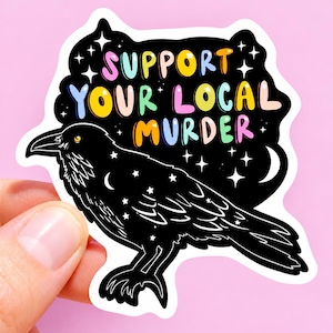 May include: A die-cut sticker featuring a black raven with white star and crescent moon patterns on its feathers. Above the bird, the text SUPPORT YOUR LOCAL MURDER is written in colorful, rounded letters against a black background with white stars.