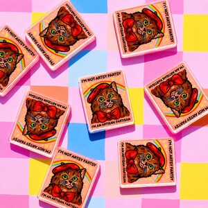 May include: Rectangular stickers featuring a cartoon cat wearing a red beret and bow tie, with a rainbow and the text "I'M NOT ARTSY FARTSY" and "I'M AN ARTISAN FARTISAN". The stickers are arranged on a pink, blue, and yellow checkered background.