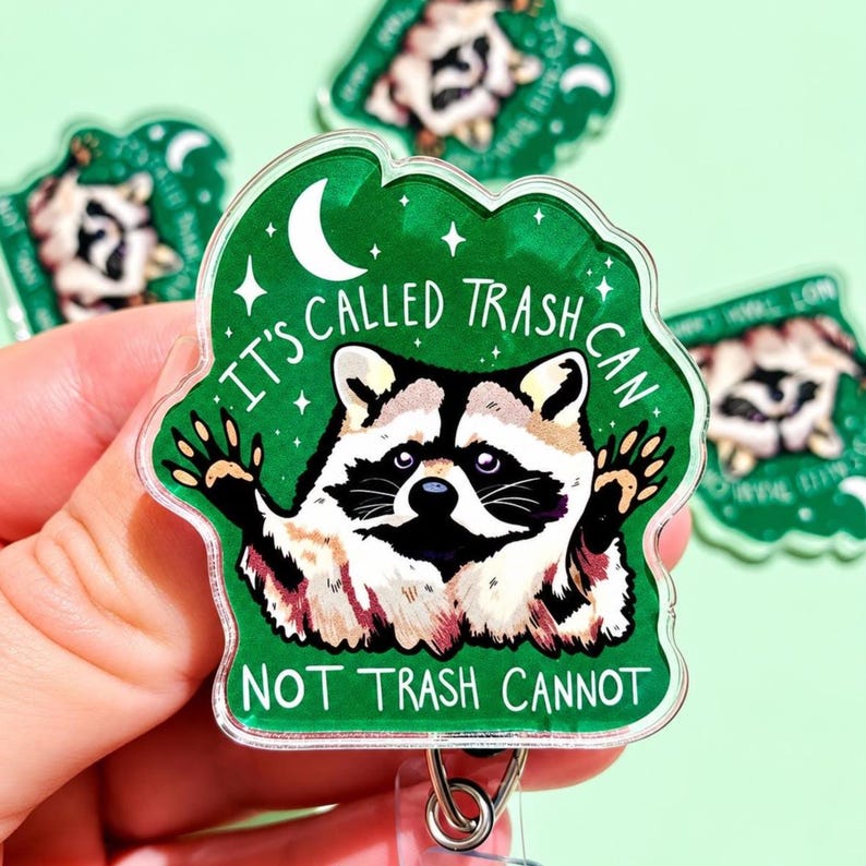 May include: A clear acrylic badge reel featuring a green background with a raccoon illustration, a white crescent moon, and stars. The text reads: IT'S CALLED TRASH CAN, NOT TRASH CANNOT. The design includes a metal clip on the bottom for attachment.