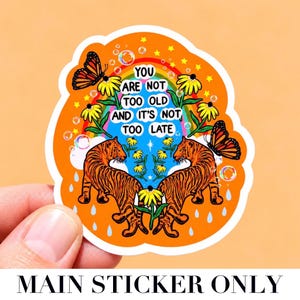 May include: A die-cut sticker with an orange background featuring two orange and black tigers facing each other. The design includes yellow flowers, monarch butterflies, a rainbow, bubbles, and the text: YOU ARE NOT TOO OLD AND IT'S NOT TOO LATE.