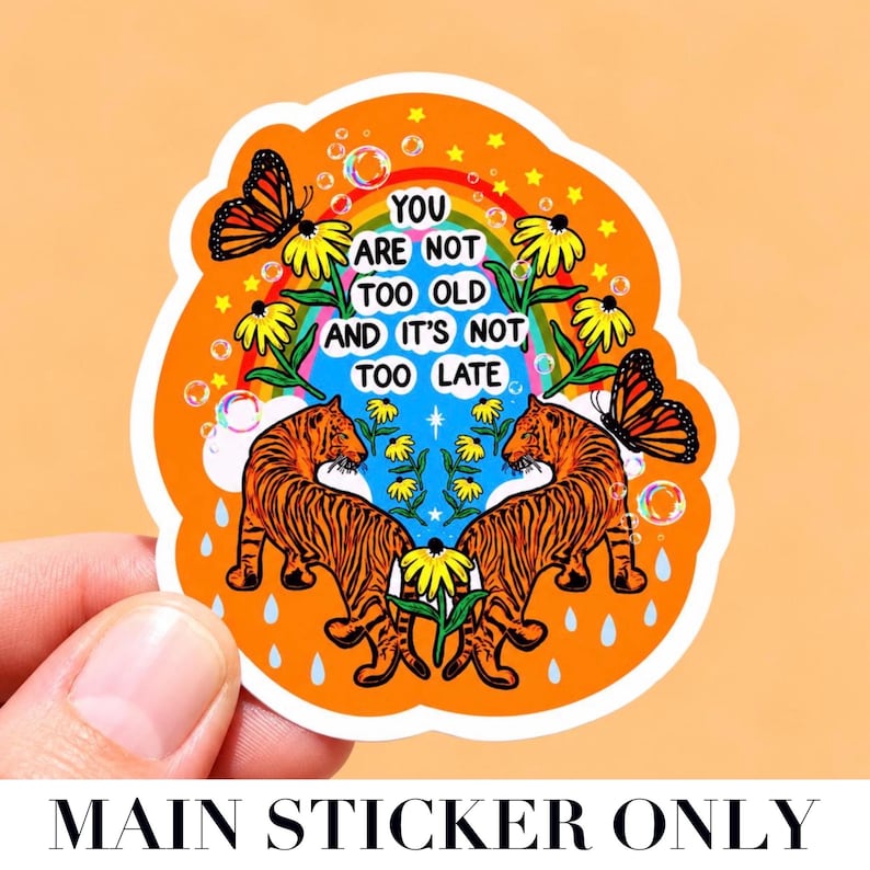 May include: A die-cut sticker with an orange background featuring two orange and black tigers facing each other. The design includes yellow flowers, monarch butterflies, a rainbow, bubbles, and the text: YOU ARE NOT TOO OLD AND IT'S NOT TOO LATE.