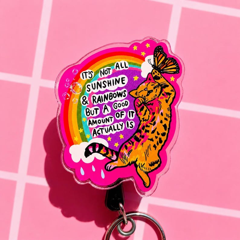 May include: A circular acrylic badge reel featuring an orange tabby cat reaching for a butterfly. The design includes a colorful rainbow, yellow stars, and the text: IT'S NOT ALL SUNSHINE & RAINBOWS BUT A GOOD AMOUNT OF IT ACTUALLY IS, set against a bright pink background.