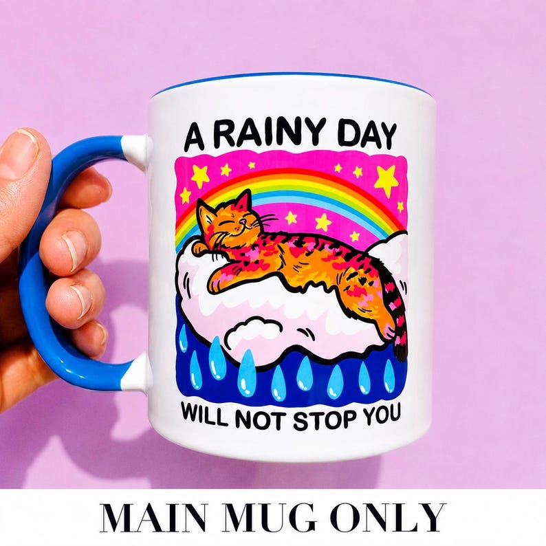 May include: A white ceramic mug with a blue handle and interior. The graphic features an orange tabby cat sleeping on a pink cloud under a rainbow and yellow stars, with blue raindrops below. The text reads A RAINY DAY WILL NOT STOP YOU.