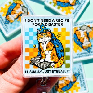 May include: A rectangular badge reel featuring an orange tabby cat wearing a blue wizard hat and cape with yellow stars. The cat sits on an open book against a light blue and yellow checkered background. Text reads: I DON'T NEED A RECIPE FOR DISASTER I USUALLY JUST EYEBALL IT.