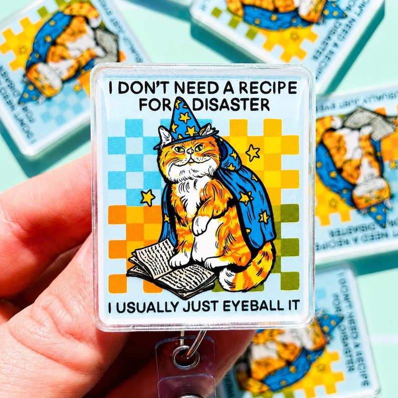 May include: A rectangular badge reel featuring an orange tabby cat wearing a blue wizard hat and cape with yellow stars. The cat sits on an open book against a light blue and yellow checkered background. Text reads: I DON'T NEED A RECIPE FOR DISASTER I USUALLY JUST EYEBALL IT.