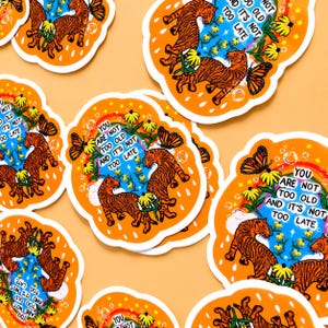 May include: Die-cut stickers featuring two orange tigers, yellow flowers, a butterfly, and a rainbow against a blue sky. The text reads, "YOU ARE NOT TOO OLD AND IT'S NOT TOO LATE." The stickers have an orange border with white raindrop shapes.